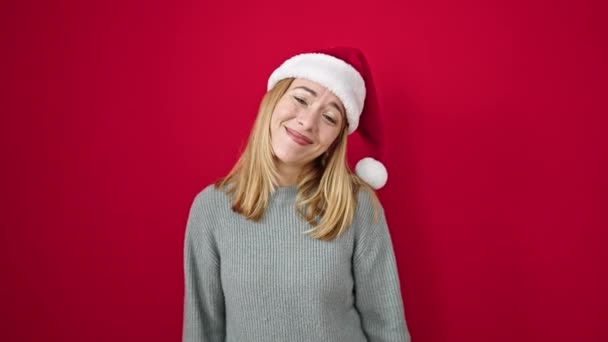 Mladá blondýna žena usmívá sebevědomě nosí vánoční klobouk přes izolované červené pozadí - Záběry, video