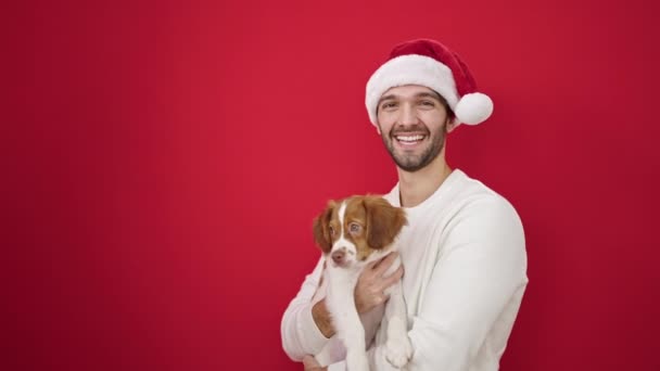 Δύο άνδρες ζευγάρι φορώντας χριστουγεννιάτικο καπέλο αγκαλιάζει το σκυλί φιλιά πάνω από απομονωμένο κόκκινο φόντο - Πλάνα, βίντεο