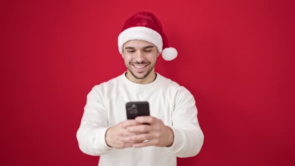 Junger hispanischer Mann trägt Weihnachtsmütze mit Smartphone vor isoliertem roten Hintergrund - Filmmaterial, Video