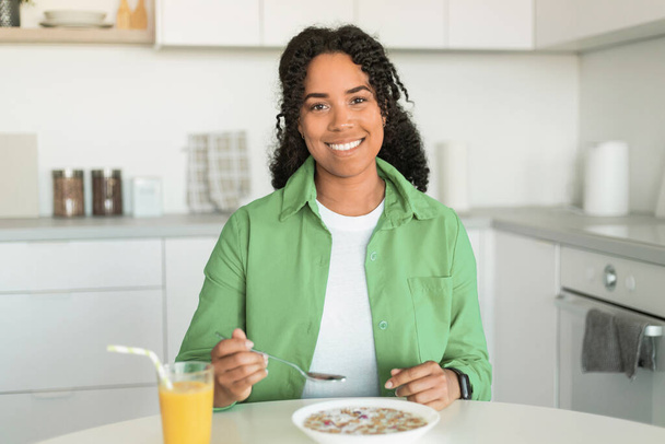 Vrolijke zwarte dame genieten van het ontbijt, zitten met bord ontbijtgranen voor haar en een glas sinaasappelsap, genieten van maaltijd en evenwichtige voeding in gezellige keuken thuis. Voeding en welzijn - Foto, afbeelding