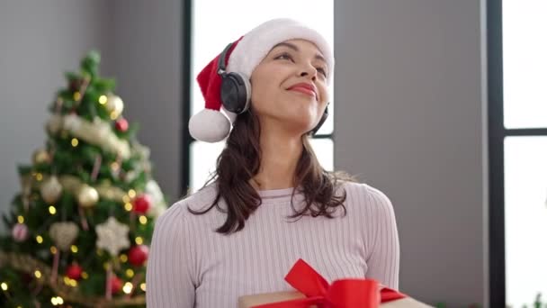 Νεαρή όμορφη Ισπανίδα που χορεύει δίπλα στο χριστουγεννιάτικο δέντρο φορώντας ακουστικά στο σπίτι - Πλάνα, βίντεο