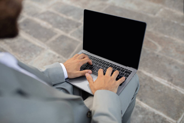 Onherkenbare zakenman werkt op laptop computer met blanco scherm buiten, mannelijke ondernemer in pak werkt online, zittend op trappen in stedelijk gebied, browsen website, mockup - Foto, afbeelding