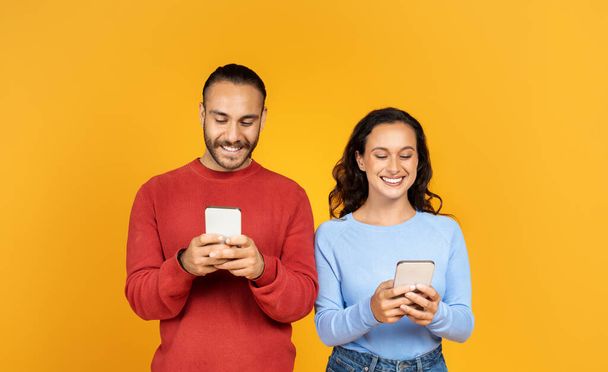 Portrait of cheerful couple holding their smartphones and playing game or chatting online. Happy millennial man and woman using gadgets, isolated on yellow studio wall - Photo, Image