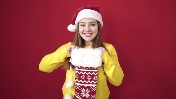 Молодая блондинка улыбается уверенно держа рождественский носок на изолированном красном фоне - Кадры, видео