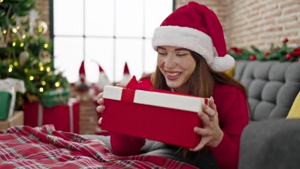 Молода іспаномовна жінка в різдвяному капелюсі розпаковує подарунок з здивованим обличчям вдома - Кадри, відео