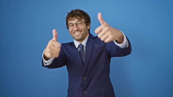 Cheerful young man in business suit celebrates success with a winning thumbs-up gesture, smiling, radiating positivity. captured solo against crisp blue isolated background. - Footage, Video
