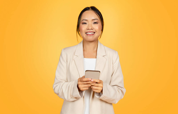 Frohe chinesische Geschäftsfrau im Anzug, die geschäftlich auf dem Smartphone tippt, plaudert, isoliert auf orangefarbenem Studiohintergrund. Work App, Anzeige und Angebot, Lifestyle mit Technologie - Foto, Bild
