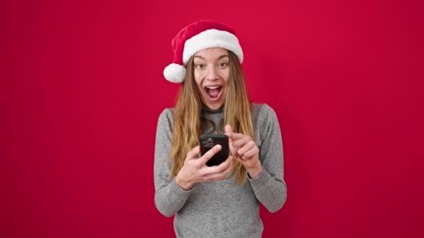 Giovane donna caucasica indossa cappello di Natale utilizzando smartphone facendo gesto ok su sfondo rosso isolato - Filmati, video
