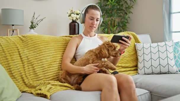 Junge kaukasische Frau mit Hund sitzt auf dem Sofa und sieht sich ein Video mit Kopfhörern zu Hause an - Filmmaterial, Video