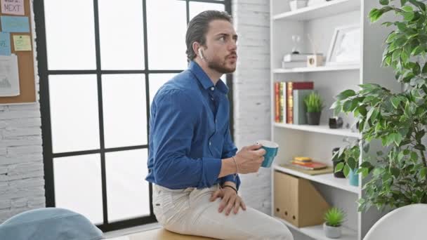 Trabajador de negocios hispano joven con auriculares bebiendo café en la oficina - Metraje, vídeo