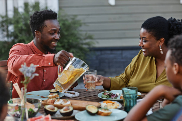 Mosolygó fekete férfi portréja, aki limonádét önt az üvegre a családi vacsora alatt a szabadban. - Fotó, kép