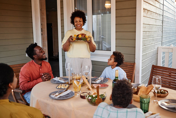 Portré idősebb fekete nő hozza az ételt az asztalra, miközben élvezi a családi összejövetel szabadban - Fotó, kép