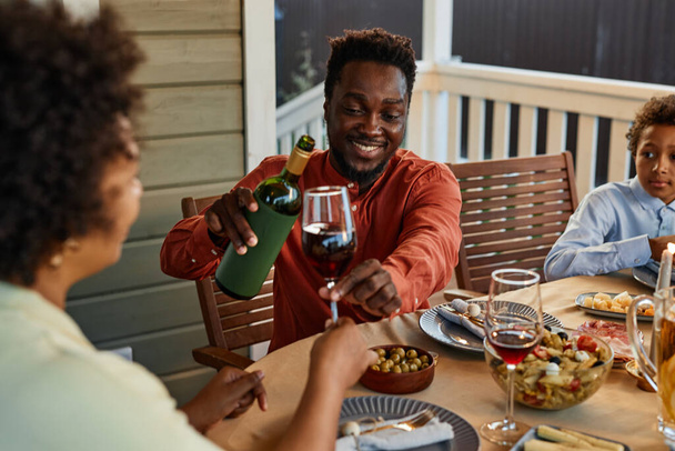 Porträt eines lächelnden schwarzen Mannes, der Wein in Gläser gießt, während er seine Dinnerparty im Freien genießt - Foto, Bild