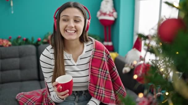 Nuori kaunis latino nainen kuuntelee musiikkia juo kahvia juhlii joulua kotona - Materiaali, video