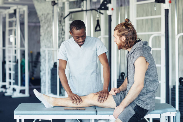Africano médico fazendo massagem na perna para paciente durante sua reabilitação no health club - Foto, Imagem