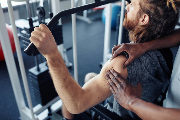 Junger Patient trainiert seinen verletzten Arm an Trainingsgeräten im Fitnessstudio, während Therapeut ihn massiert - Foto, Bild