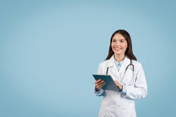 Nuori eurooppalainen naislääkäri valkoisessa lääketieteellisessä univormussa, digitaalinen tabletti kädessään ja katsoen kopiotilaa sinistä taustaa vasten lääketieteellisiä mainoksia varten, banneri - Valokuva, kuva
