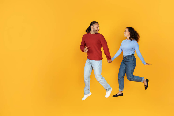 Joyful millennial couple holding hands and running towards copy space, happy young man and woman in love enjoying romantic date, isolated on yellow studio background, full length - Photo, Image