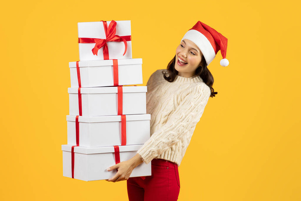 Happy young woman with stack of wrapped Christmas presents boxes advertising holiday sales season, posing wearing Santa hat on yellow studio background. Winter sale banner - Photo, Image
