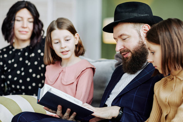 Zijaanzicht portret van orthodoxe joodse man lezen boek aan kinderen in huis instelling - Foto, afbeelding