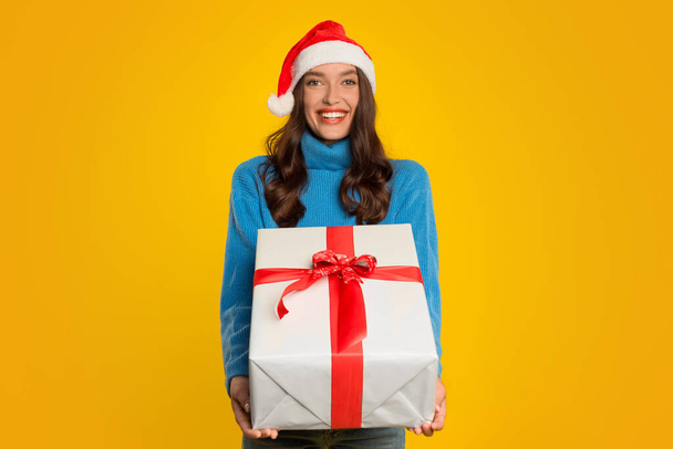 Счастливая молодая женщина весело позирует с обернутой подарочной коробкой, предлагая Рождество подарок на камеру, на желтом фоне студии. Леди в Санта-шляпе рекламирует зимние предложения и поздравления - Фото, изображение