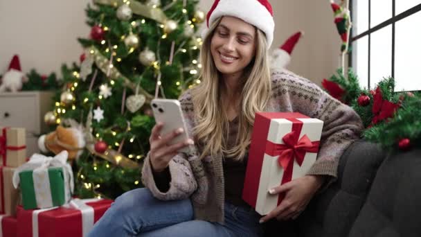 Молода блондинка за допомогою смартфона тримає подарунок, сидячи на ялинці вдома - Кадри, відео