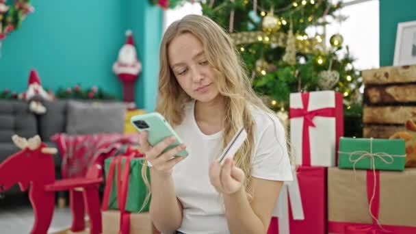 Nuori blondi nainen ostoksilla älypuhelimella ja luottokortilla juhlii joulua kotona - Materiaali, video