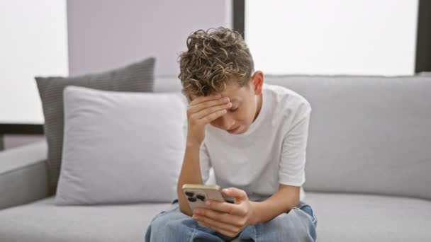 Adorable blond boy, stressed and upset, sitting on sofa at home using smartphone. worried child typing a message, showing serious expression amidst technology stress. - Footage, Video