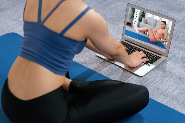 Asian woman in sportswear doing yoga exercise on fitness mat as her home workout training routine. Healthy body care lifestyle woman watching online yoga video on laptop. Vigorous - Photo, Image