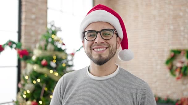 Ισπανόφωνος άντρας χαμογελάει με αυτοπεποίθηση φορώντας χριστουγεννιάτικο καπέλο στο σπίτι - Πλάνα, βίντεο