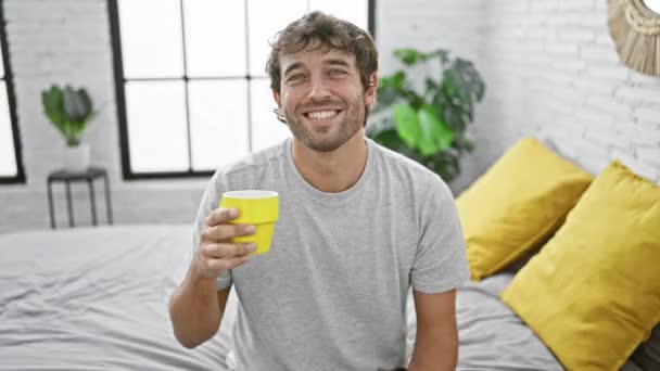 Iloinen nuori mies, siemaillen aamukahvia sängyssä, innokkaasti osoittaa ajatus kaikki hymyilee ja paistaa hampaat mukavasti kotiinsa makuuhuoneessa - Materiaali, video