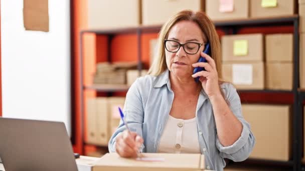 Mujer hispana de mediana edad comercio electrónico trabajador de negocios escribiendo en el paquete de hablar en el teléfono inteligente en la oficina - Imágenes, Vídeo
