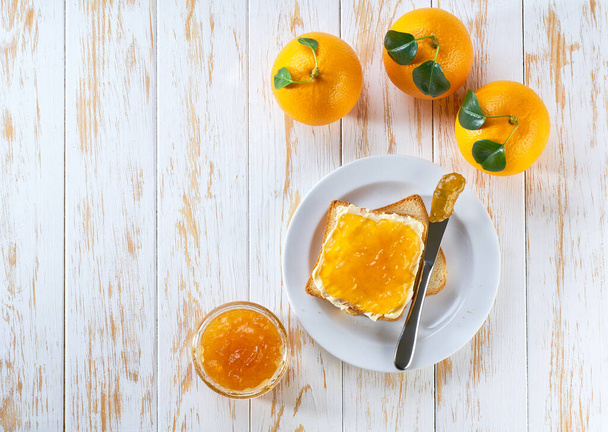 toast met sinaasappeljam, pot jam, mes op een lichte houten tafel. zicht van bovenaf - Foto, afbeelding