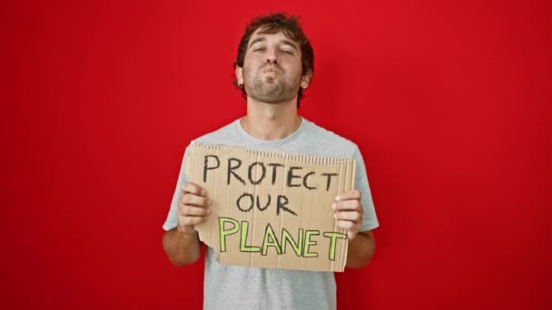 Havayla şişirilmiş, yanakları şişirilmiş genç bir sarışının komik ifadesi. Elinde izole edilmiş kırmızı bir arka planda "gezegenimizi koruyun" afişi tutarken. - Video, Çekim