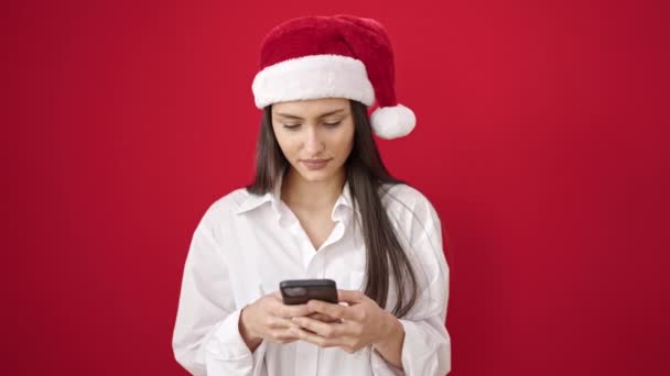 Jeune belle femme hispanique portant un chapeau de Noël en utilisant un smartphone sur fond rouge isolé - Séquence, vidéo