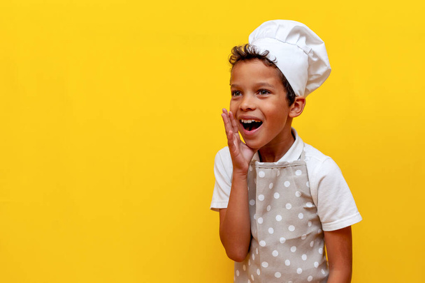 niño afroamericano en uniforme y sombrero de chef anuncia en voz alta y grita sobre fondo amarillo aislado, niño adolescente en delantal habla información con la boca abierta - Foto, Imagen