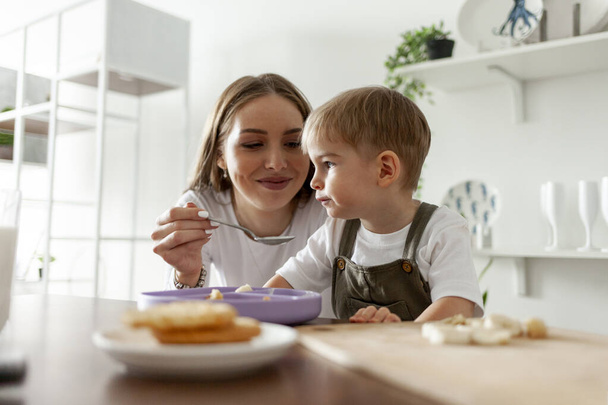 young mother feeds her little son with spoon in the kitchen at home, 2-year-old boy has breakfast with his parent, woman gives the child something to eat and smiles - Photo, Image