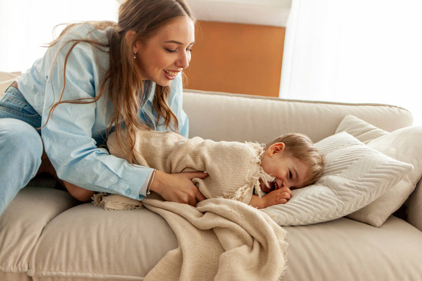 jonge moeder speelt met haar kleine zoon thuis op de bank en bedekt hem met deken, 2-jarige jongen lacht en heeft plezier met zijn ouders, vrouw zet het kind naar bed - Foto, afbeelding
