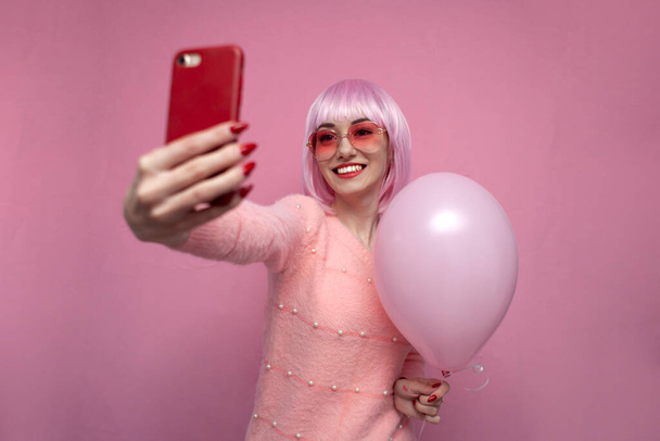Nuori tyttö, jolla on vaaleanpunaiset hiukset, ottaa selfien älypuhelimella, nainen, jolla on lyhyt värillinen kampaus, ottaa valokuvan vaaleanpunaisella ilmapallolla. - Valokuva, kuva