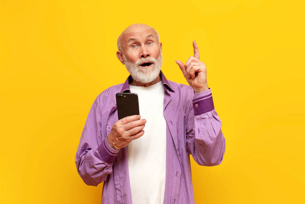 Mam pomysł. stary łysy dziadek w fioletowej koszuli używa smartfona i podnosi palec na żółtym, odizolowanym tle, emeryt starzec z planami telefonów komórkowych - Zdjęcie, obraz