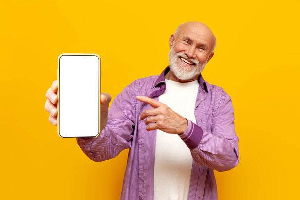 old bald grandfather in purple shirt shows blank smartphone screen on yellow isolated background, elderly pensioner advertises and points to the mobile display - Photo, Image