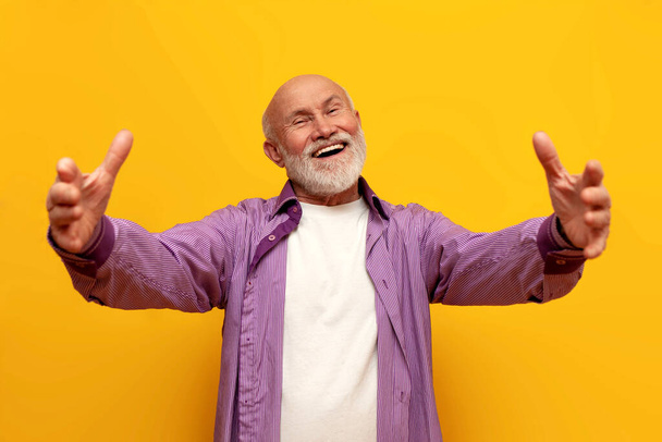 stary łysy dziadek w fioletowej koszuli ciągnie ręce do przodu i przytula się do żółtego, odizolowanego tła, emeryt pokazuje mile widziany gest i trzyma przestrzeń do kopiowania - Zdjęcie, obraz