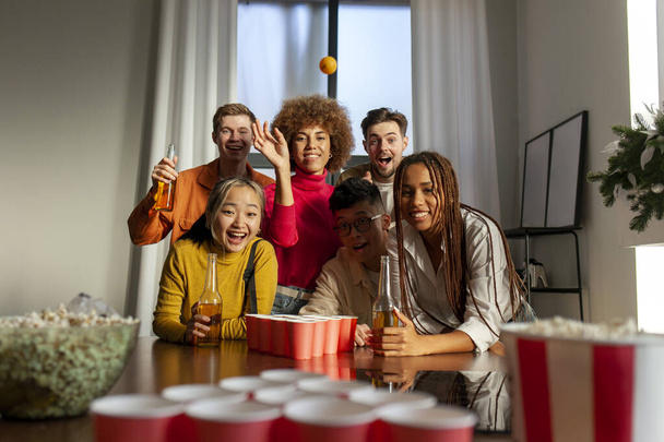 monirotuinen ryhmä nuoria ystäviä pelaamassa olutpingistä juhlissa juomassa olutta ja pitämässä hauskaa kotona, opiskelijat juomassa alkoholia ja juhlimassa voittoa - Valokuva, kuva