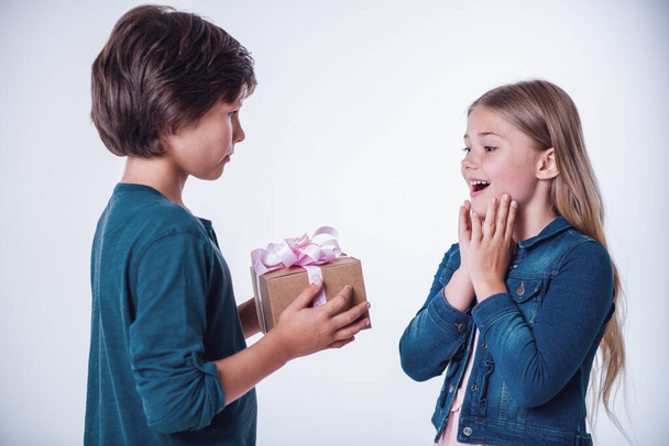 Очаровательная маленькая девочка улыбается и удивляется, когда красивый мальчик дарит ей подарочную коробку.
 - Фото, изображение