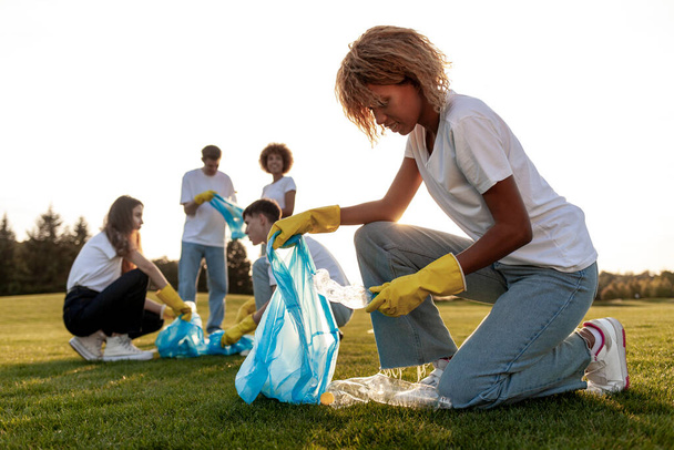 groupe de personnes multiraciales dans des gants et avec des sacs à ordures enlever le plastique et les ordures dans le parc, équipe de bénévoles travaillent et collectent des bouteilles en plastique, les gens se soucient de l'environnement, planète verte - Photo, image
