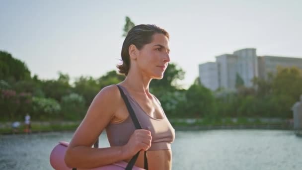 Fitness lady walking sunshine alley closeup. Tanned slim woman carrying rubber carpet looking camera at green park lake portrait. Fit sportswoman going to practicing yoga at summer. Healthy lifestyle  - Footage, Video