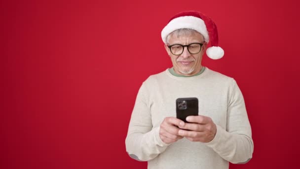 Середньовічний сіро-волохатий чоловік у різдвяному капелюсі за допомогою смартфона над ізольованим червоним тлом - Кадри, відео