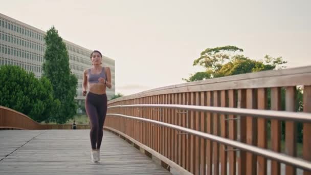 Zmotywowany sportowiec biegający porannym mostem. Muskularna kobieta ćwicząca cardio jogging w odzieży sportowej. Brunetka profesjonalny biegacz sprinting trening w parku zbliżenie. Koncepcja wytrzymałości wytrzymałościowej - Materiał filmowy, wideo