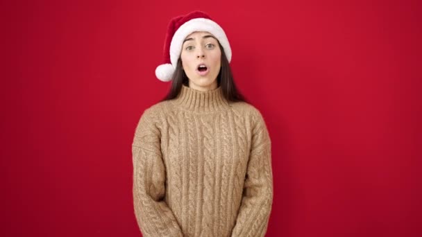Молода красива іспаномовна жінка посміхається впевнено носить різдвяний капелюх над ізольованим червоним тлом - Кадри, відео