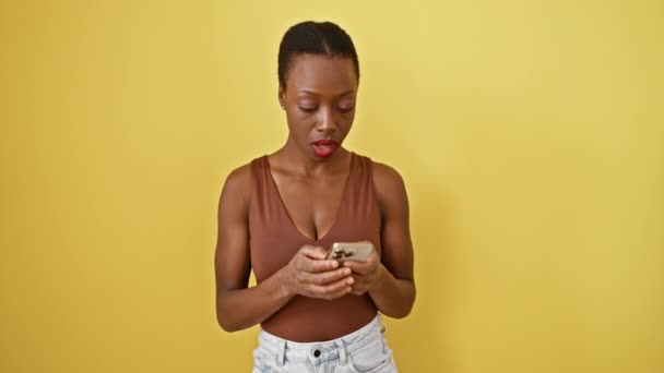 Astonished african american woman's funny shocked face with open mouth, holding smartphone. nervously texting, she's scared, amazed, in disbelief against isolated yellow backdrop. - Footage, Video